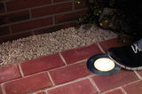 Round stainless steel recessed floor light on red brick pavement with black sneaker nearby.