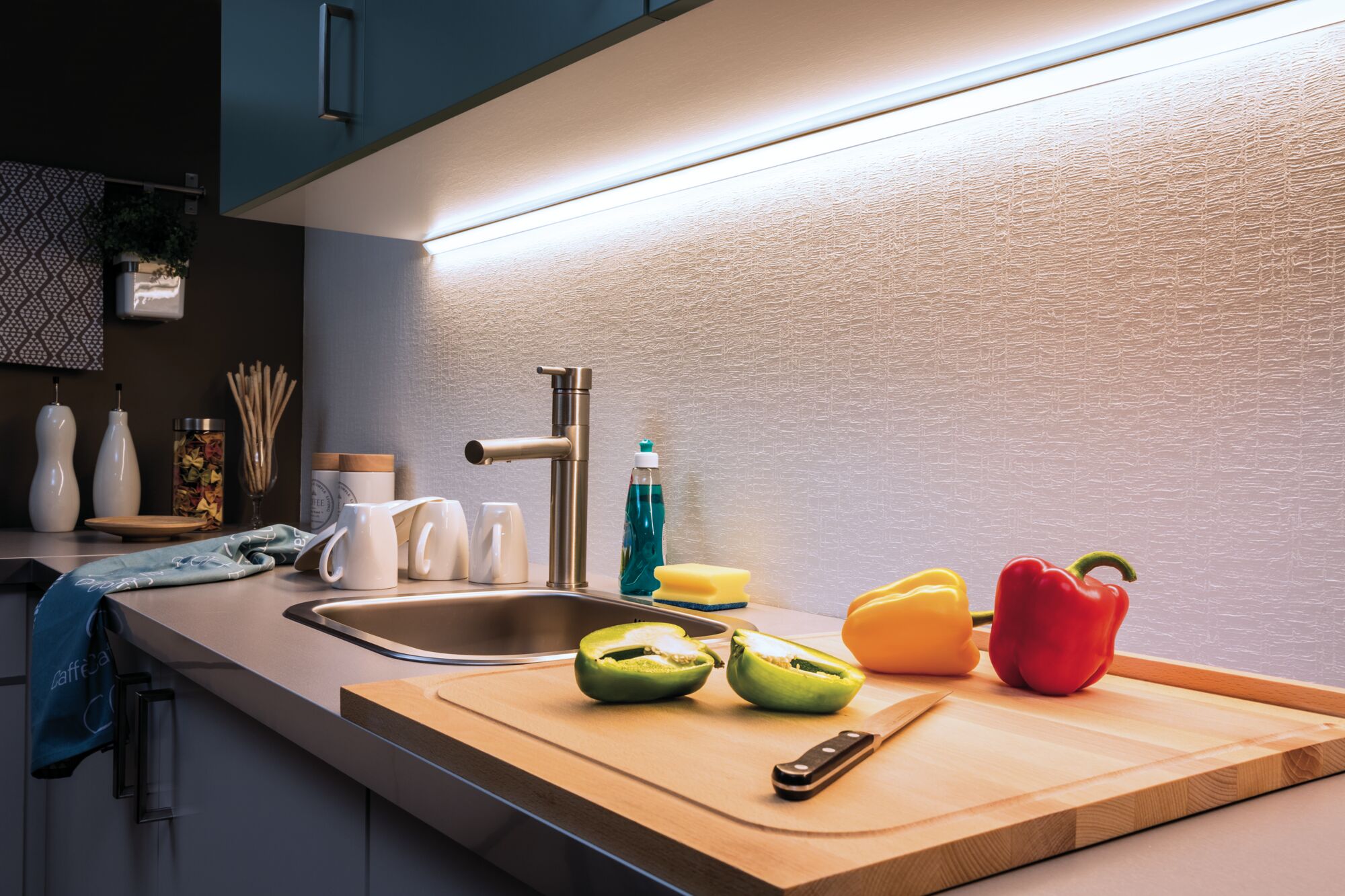 Plan de travail de cuisine moderne avec évier en acier inoxydable, planche à découper en bois et poivrons colorés sous lumière LED sous meuble