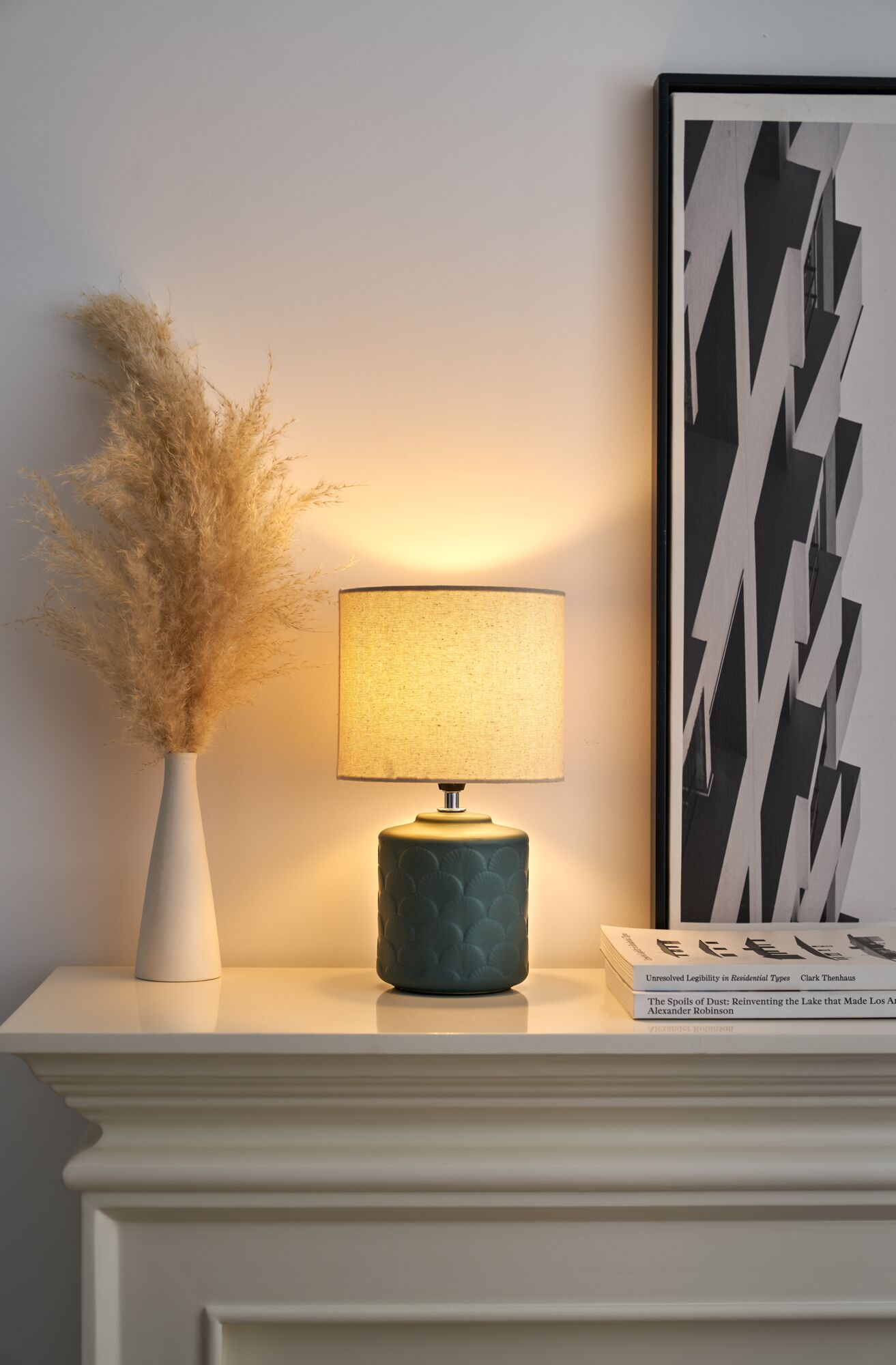 Tischlampe mit grünem Keramikfuß und beigem Stoffschirm auf weißem Kaminsims mit Deko und Bild.