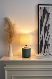 Green ceramic table lamp with beige fabric shade on white mantelpiece next to pampas grass vase