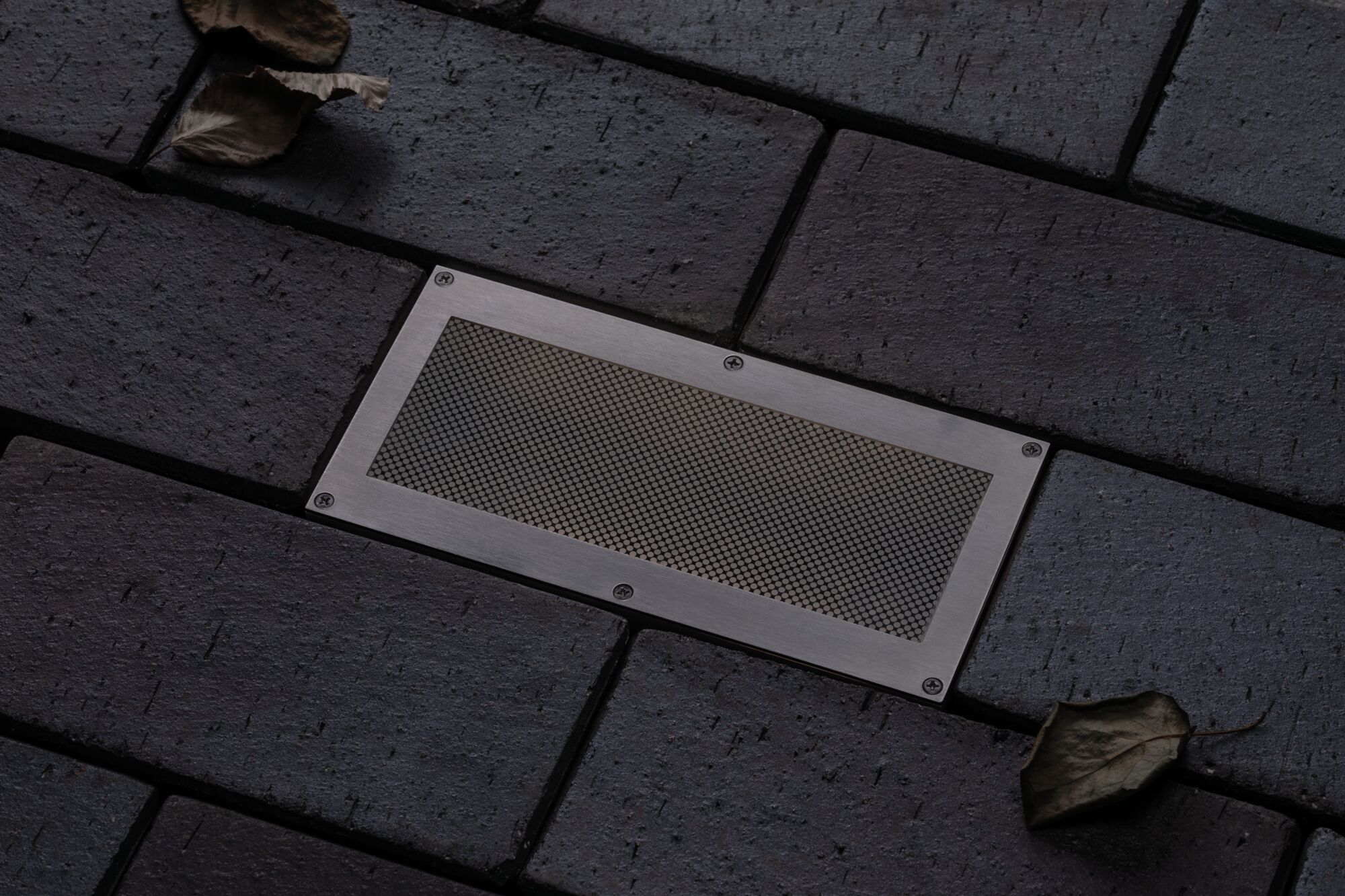 Rectangular stainless steel floor light with mesh insert in dark pavement for outdoor lighting.