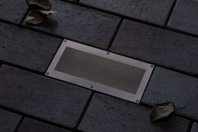 Rektangulært gulvlys i rustfrit stål med netindsats i mørk belægning til udendørs belysning.