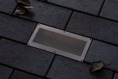 Spot de sol rectangulaire en acier inoxydable avec grille intégré dans pavé foncé pour éclairage extérieur.