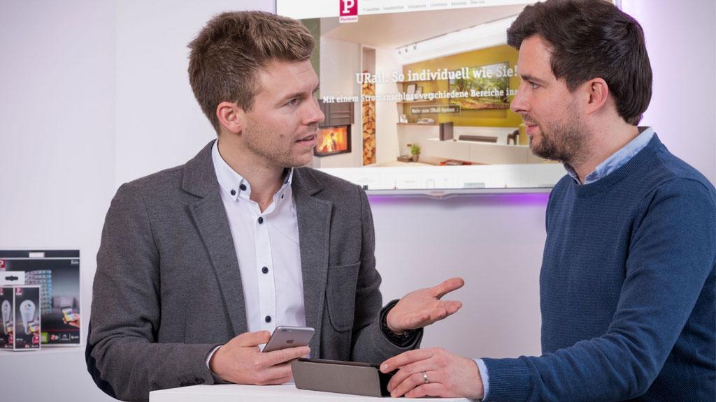 Zwei Männer im Gespräch vor Bildschirm mit Beleuchtungslösungen, Beratungssituation im Büro.