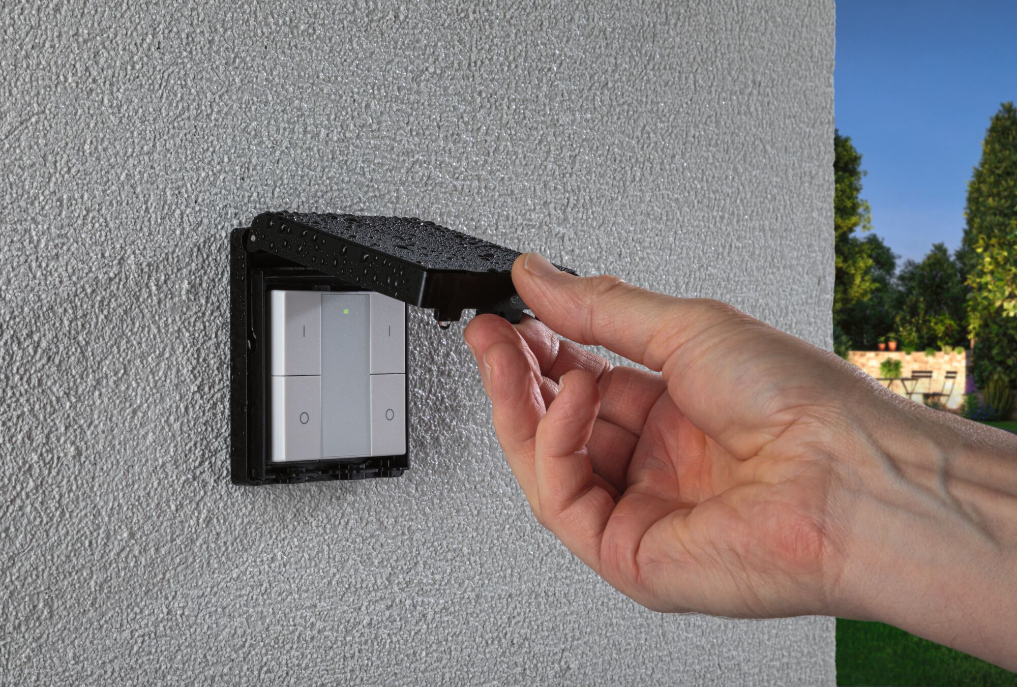 Hand lifting black rain cover with water drops over white outdoor light switch on textured wall