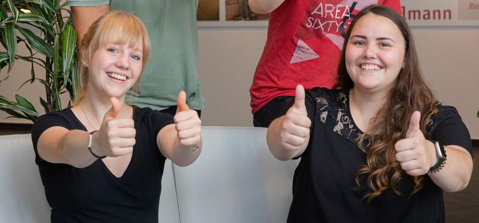 Two young women in black clothes giving thumbs up in a modern office with plants.