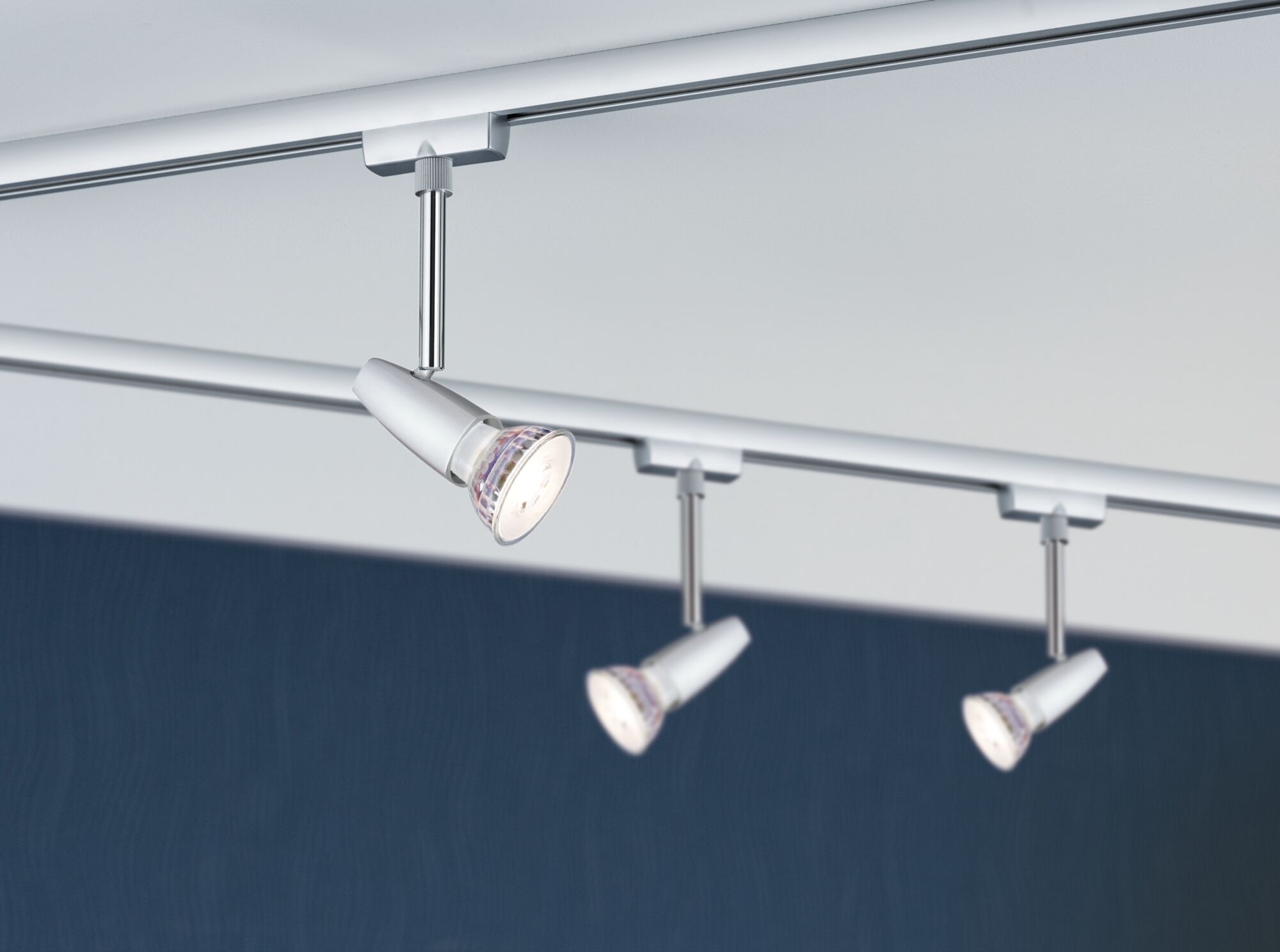 Drei weiße LED-Strahler aus Metall an Schienensystem vor dunkelblauer Wand für Akzentbeleuchtung