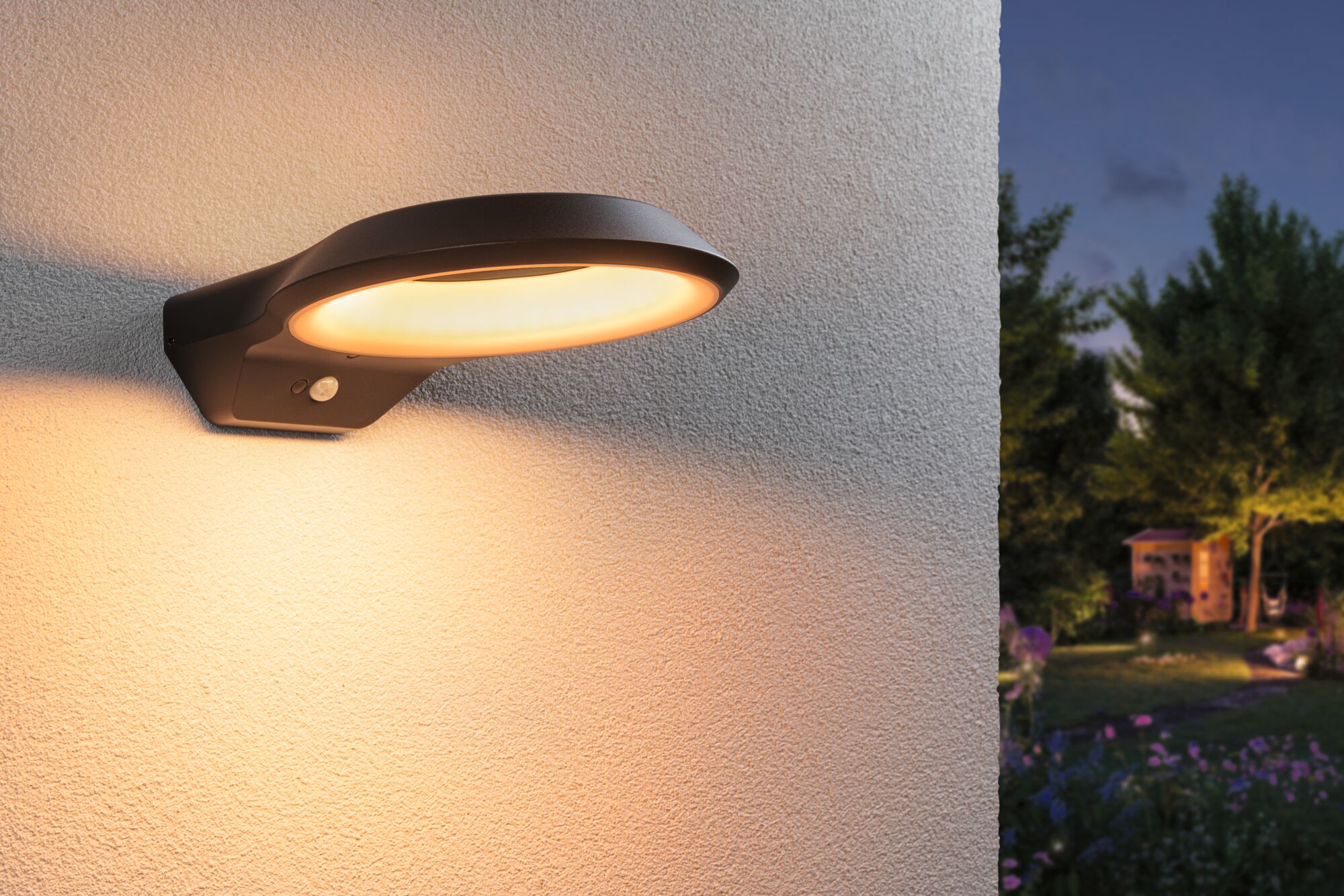 Black LED outdoor wall light with warm white glow and motion sensor mounted on house wall at dusk