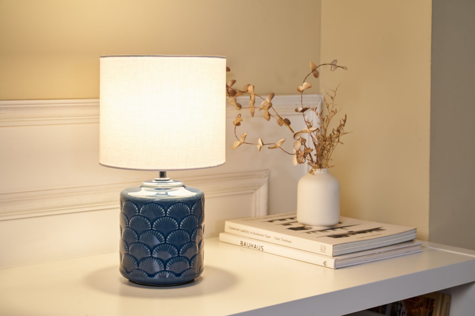 Lampe de table avec base en céramique bleue et abat-jour en tissu blanc sur table blanche avec vase et livres