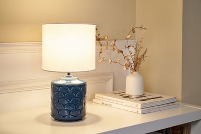 Tischlampe mit blauem Keramikfuß und weißem Stoffschirm auf weißem Tisch neben Vase und Büchern