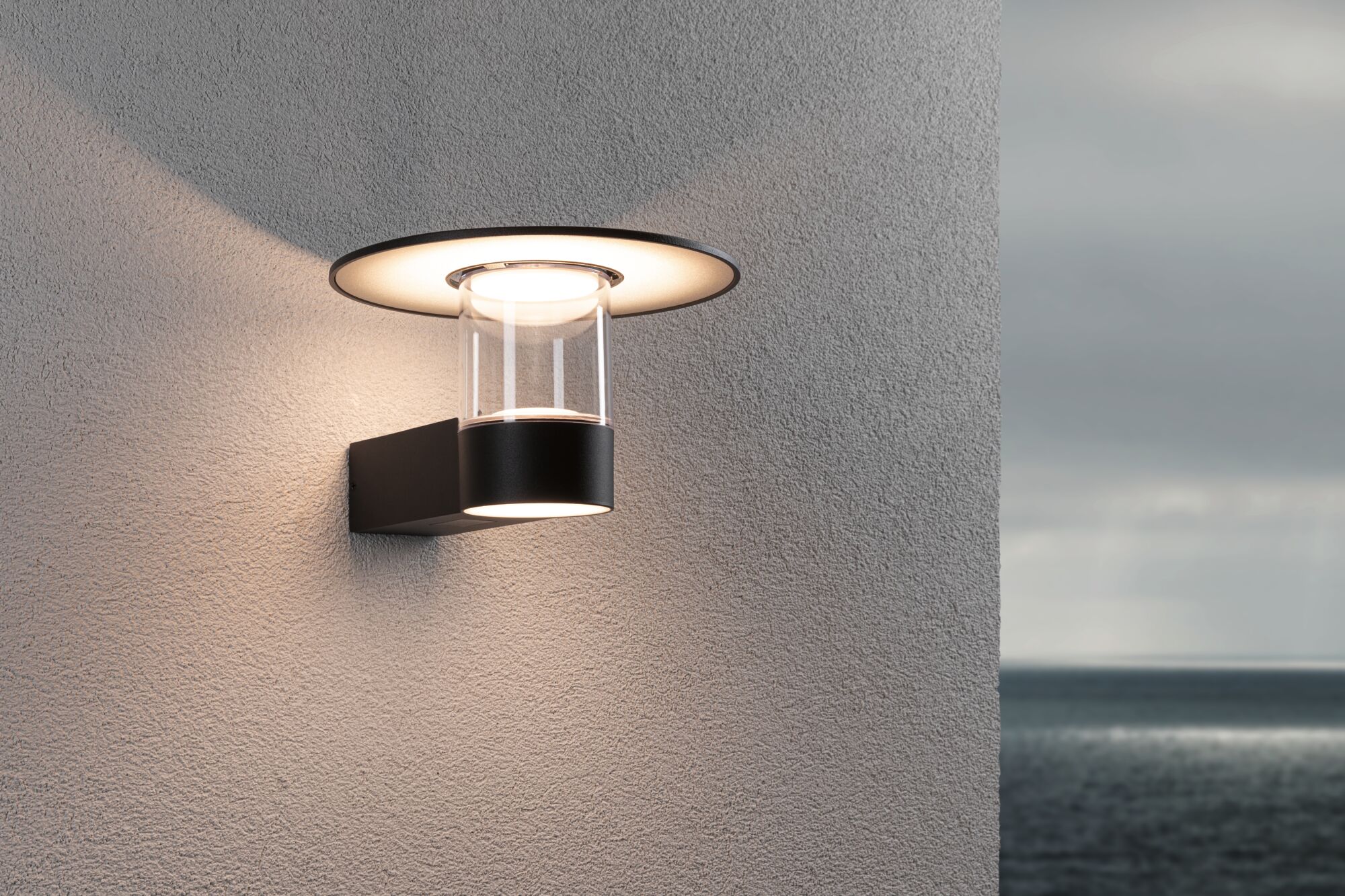 Modern black outdoor wall light made of metal and glass with warm white illumination on grey wall.
