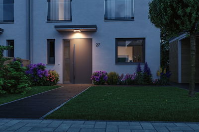 Maison moderne avec porte d'entrée grise et éclairage LED anneau blanc chaud, jardin fleuri coloré.