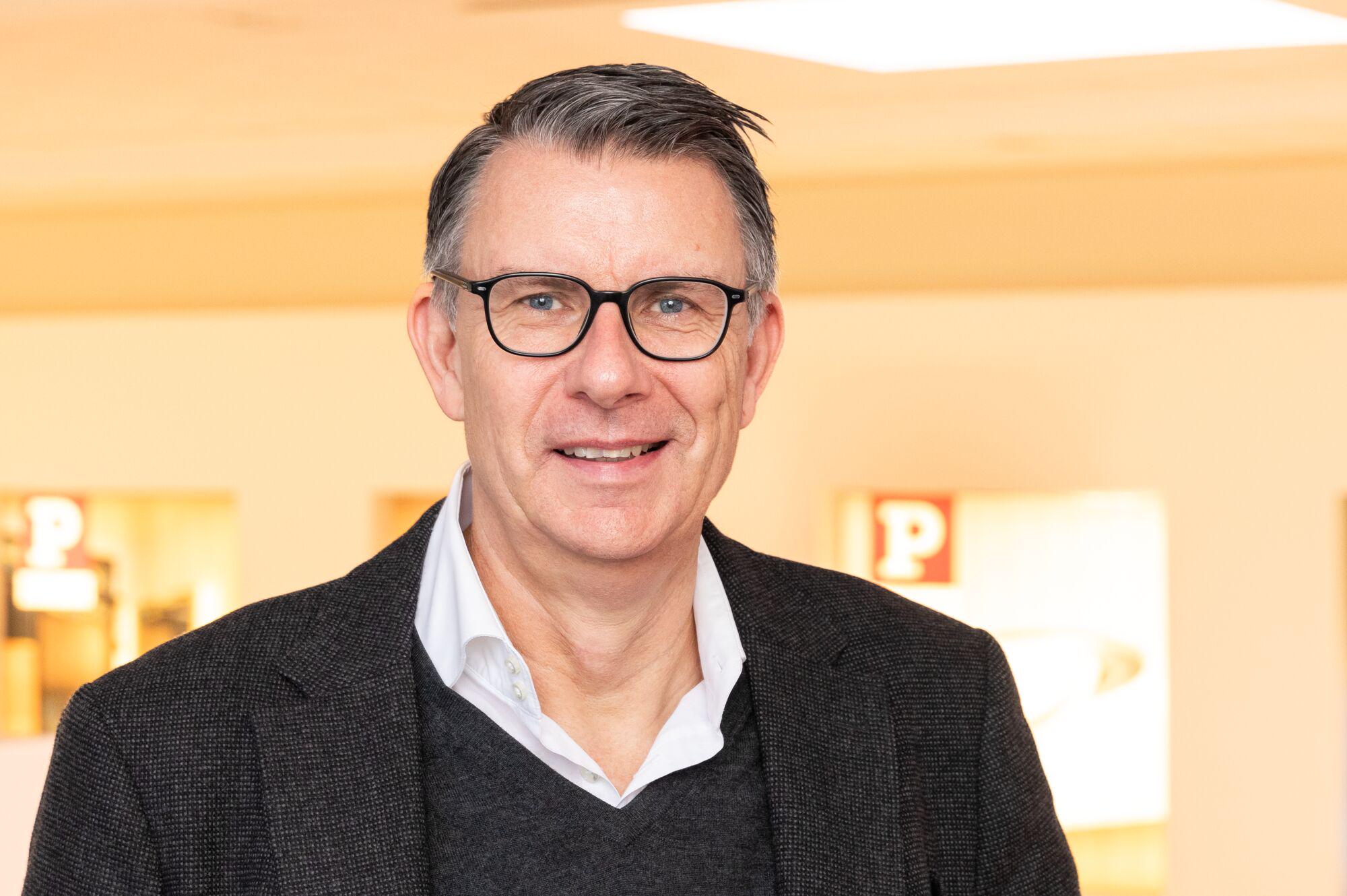 Portrait d'un homme avec lunettes et veste grise devant un fond flou et lumineux