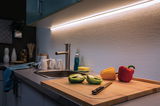 Plan de travail de cuisine moderne avec évier en acier inoxydable, planche à découper, poivrons colorés et éclairage LED sous meuble.