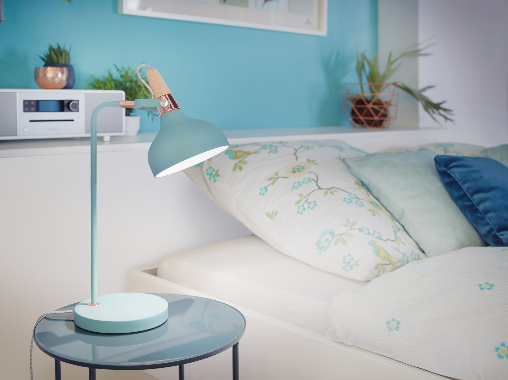 Pastel blue metal desk lamp with wooden handle on glass side table next to bed with floral bedding