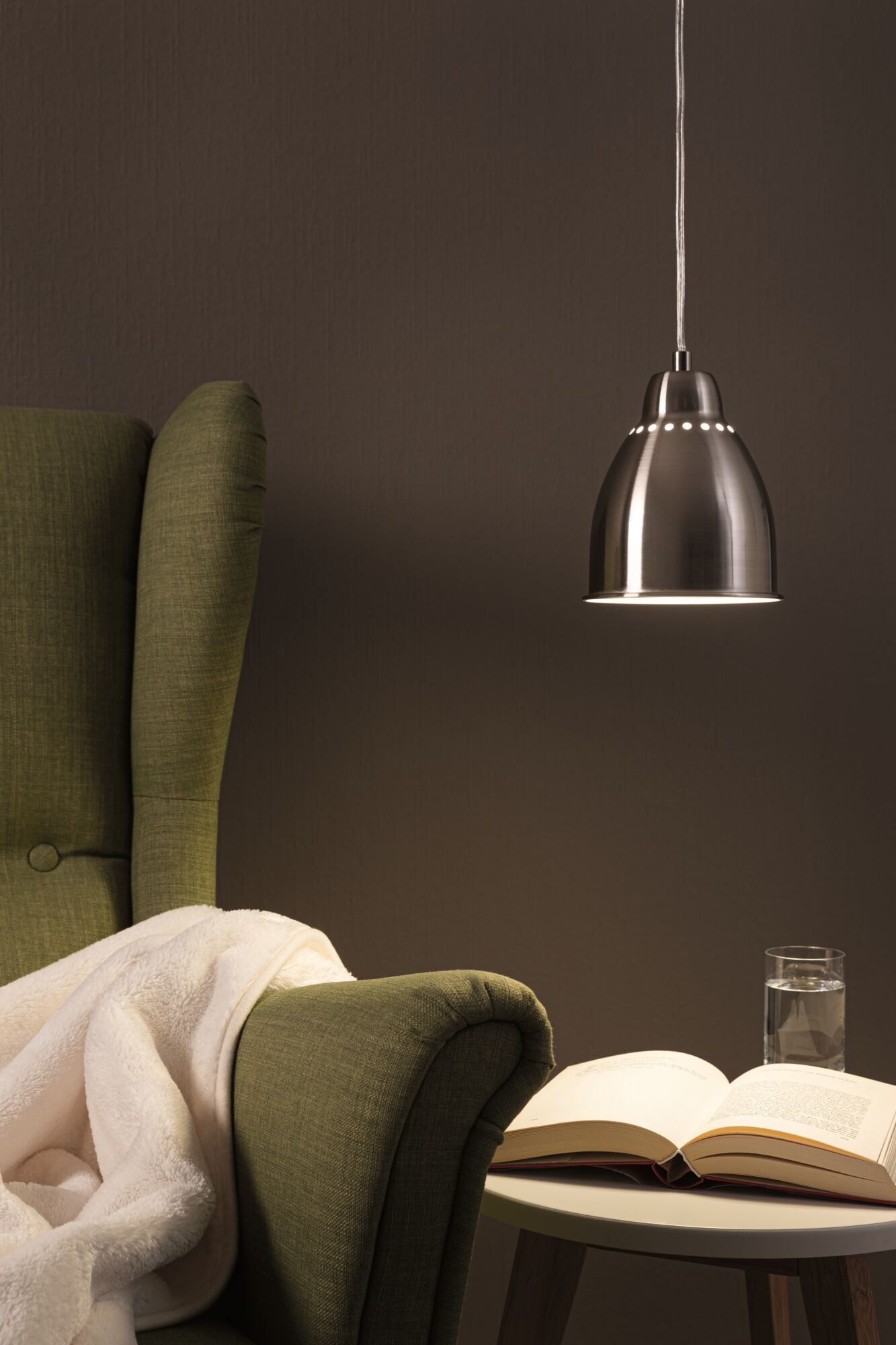 Moderner Edelstahl-Hängeleuchter mit warmweißem Licht über grünem Sessel und Lesetisch im Wohnzimmer