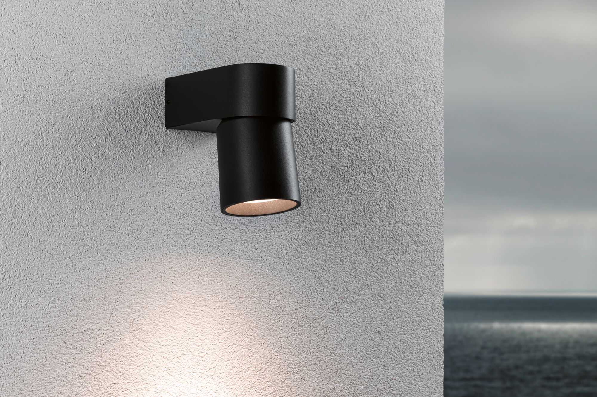 Moderner schwarzer Aluminium-Wandstrahler mit warmweißem Licht an grauer Außenwand mit Meerblick