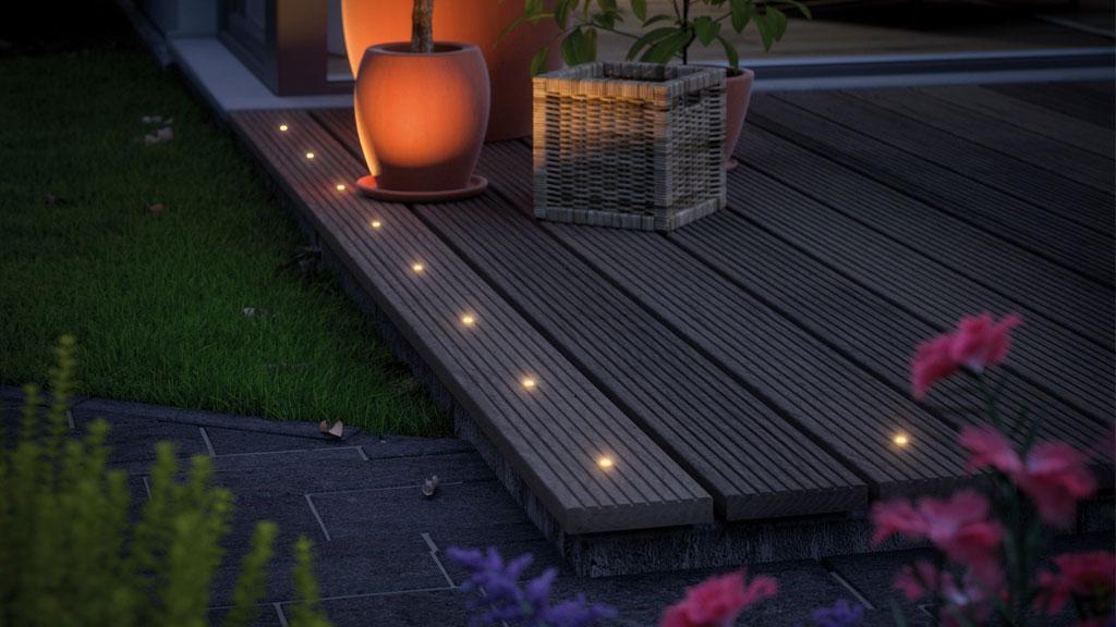 LED-Bodenlichter in warmweiß auf dunklem Holzdeck neben Pflanzen für stimmungsvolle Außenbeleuchtung.