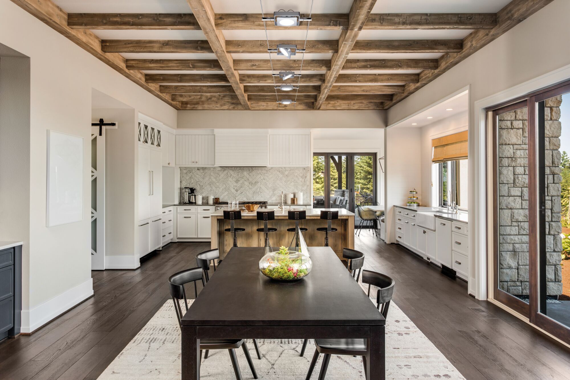 Modern kitchen with wooden ceiling beams, white cabinets, dark wood dining table with plant decor, spotlight lighting