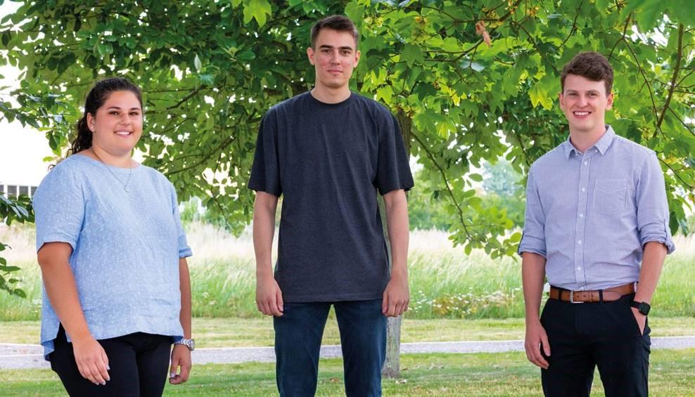 Drei junge Erwachsene stehen lächelnd im Freien vor grünem Baum, symbolisieren Ausbildung und Karriere.