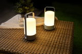 Two portable grey LED table lamps with warm white light on woven garden table surface.