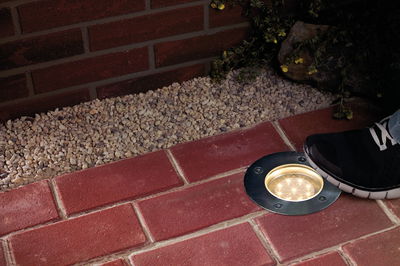 Round LED recessed floor light with stainless steel frame on red pavement with black sneaker.