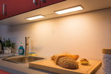 Kitchen countertop with stainless steel sink, wooden cutting board, sesame bread and muffin under warm white LED under-cabinet light