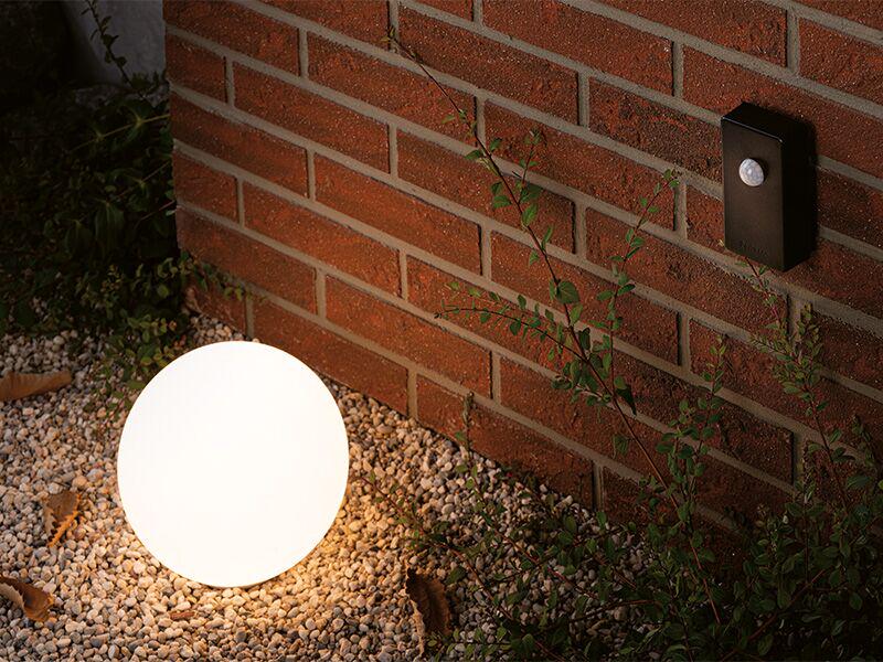 Ronde witte buitenlamp op grind bij rode bakstenen muur met zwarte bewegingssensor.