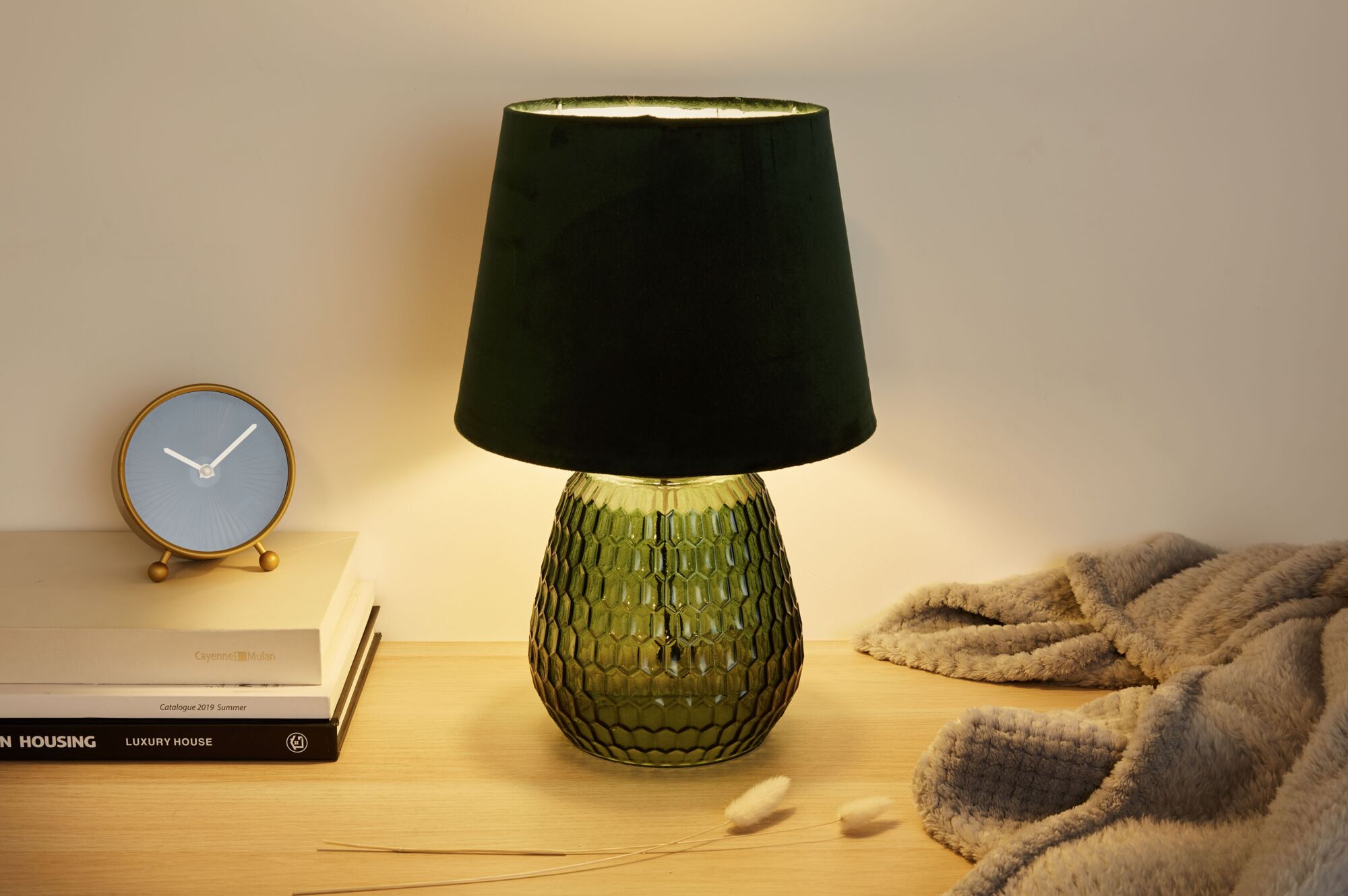 Tafellamp met groene glazen voet en zwarte stoffen kap op houten oppervlak naast klok en deken