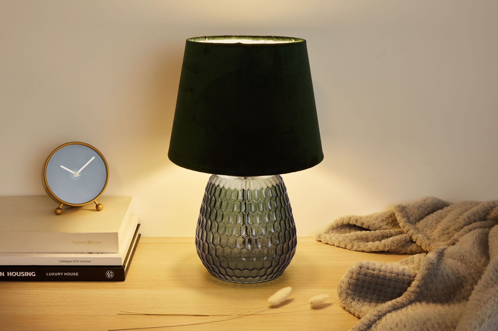 Groene glazen tafellamp met zwarte stoffen kap op houten oppervlak naast klok en deken