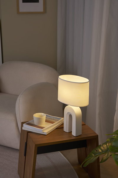Witte tafellamp met stoffen kap en boogvoet op houten tafel naast beige bank en boek