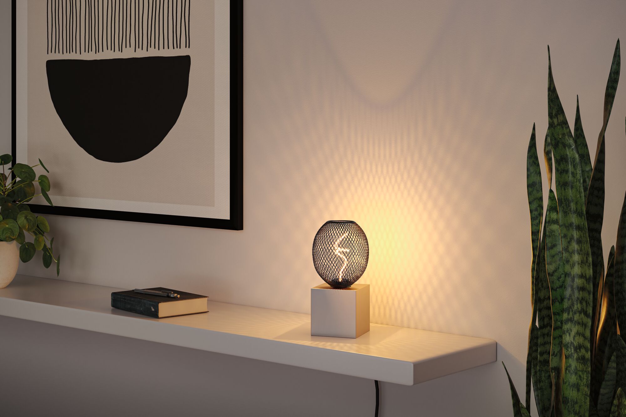 Moderne Tischleuchte mit schwarzem Metallgitter und weißem Sockel erzeugt warmes Lichtmuster an Wand.