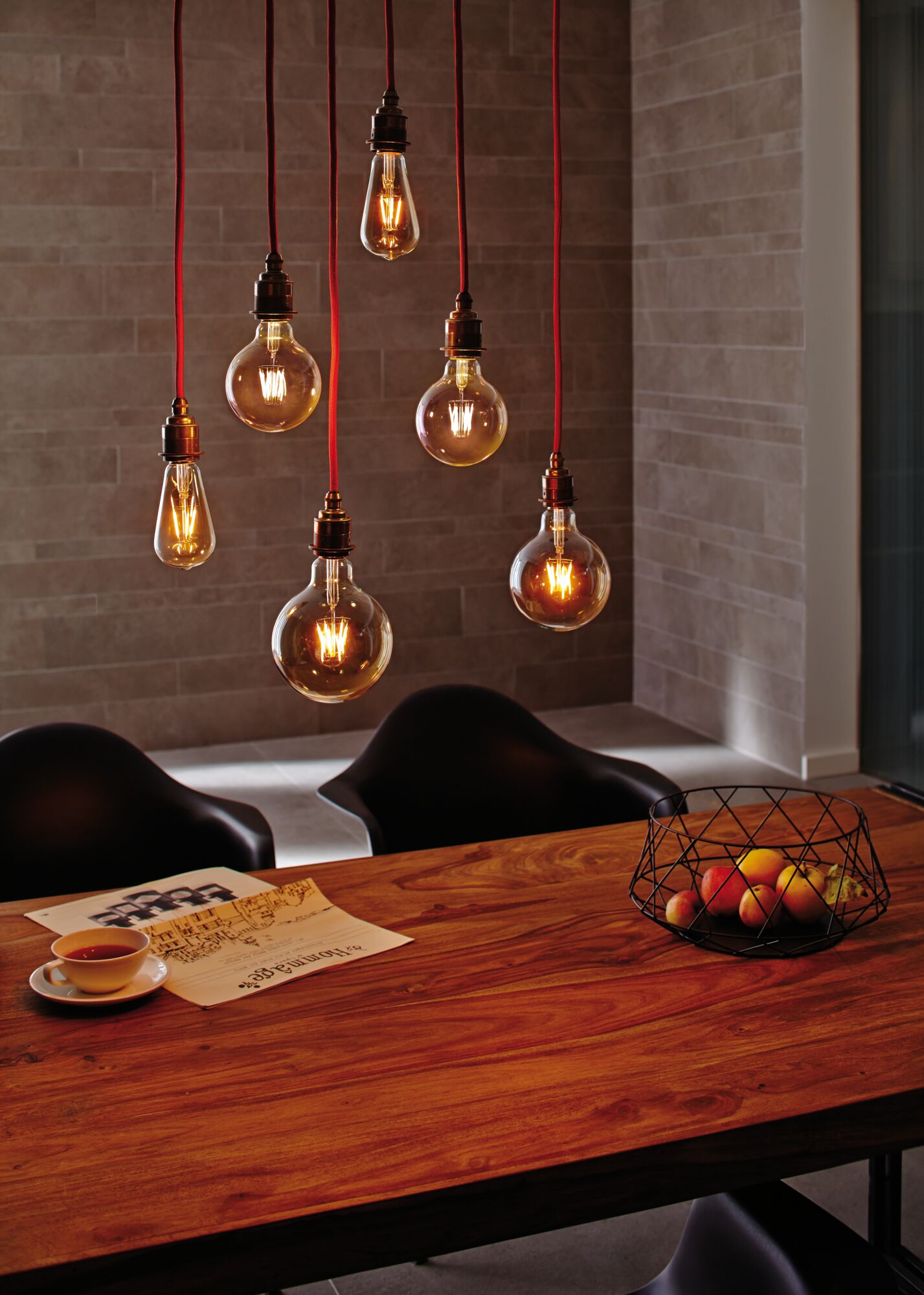 Zes hangende vintage gloeilampen met rode snoeren boven een houten tafel met fruitmand en krant.