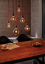 Zes hangende vintage gloeilampen met rode snoeren boven een houten tafel met fruitmand en krant.