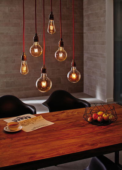 Zes hangende vintage gloeilampen met rode snoeren boven een houten tafel met fruitmand en krant.