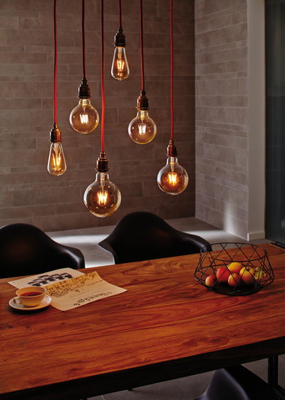 Six hanging vintage light bulbs with red cords above a wooden table with fruit basket and newspaper.
