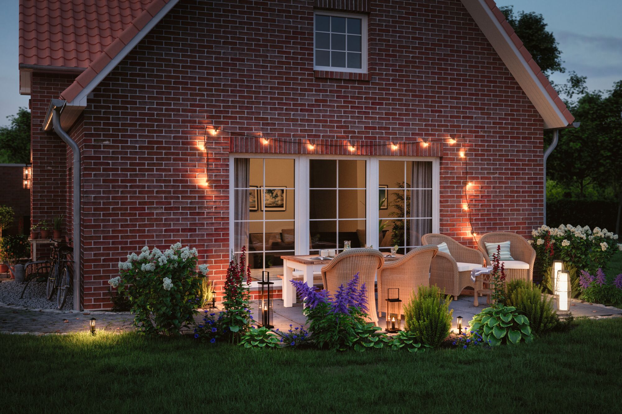 Gemütliche Terrasse mit warmweißer LED-Gartenbeleuchtung und Rattanmöbeln vor rotem Backsteinhaus.