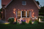 Gemütliche Terrasse mit warmweißer LED-Gartenbeleuchtung und Rattanmöbeln vor rotem Backsteinhaus.