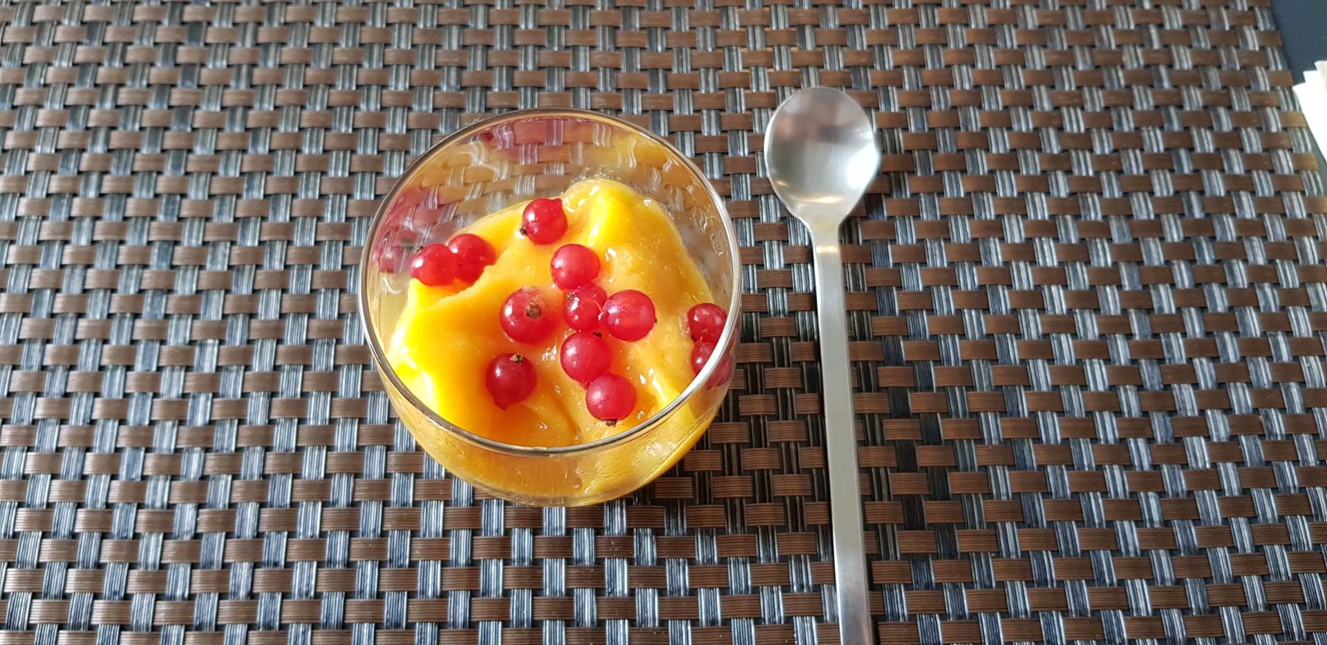 Glas mit gelbem Fruchtpüree und roten Johannisbeeren auf geflochtenem Tischset mit Metalllöffel.