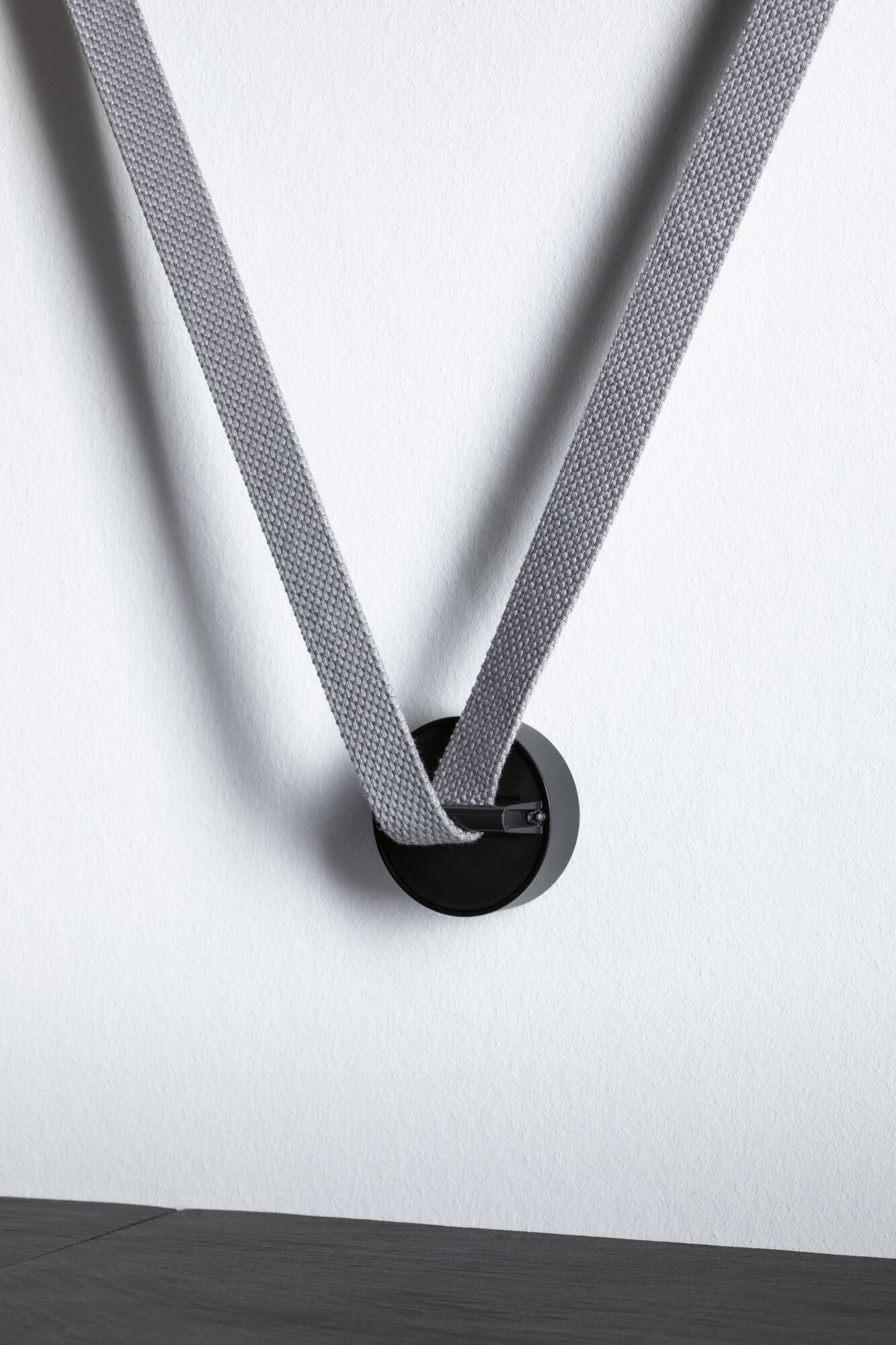 Black metal wall hook with grey fabric strap on white wall for modern interior design