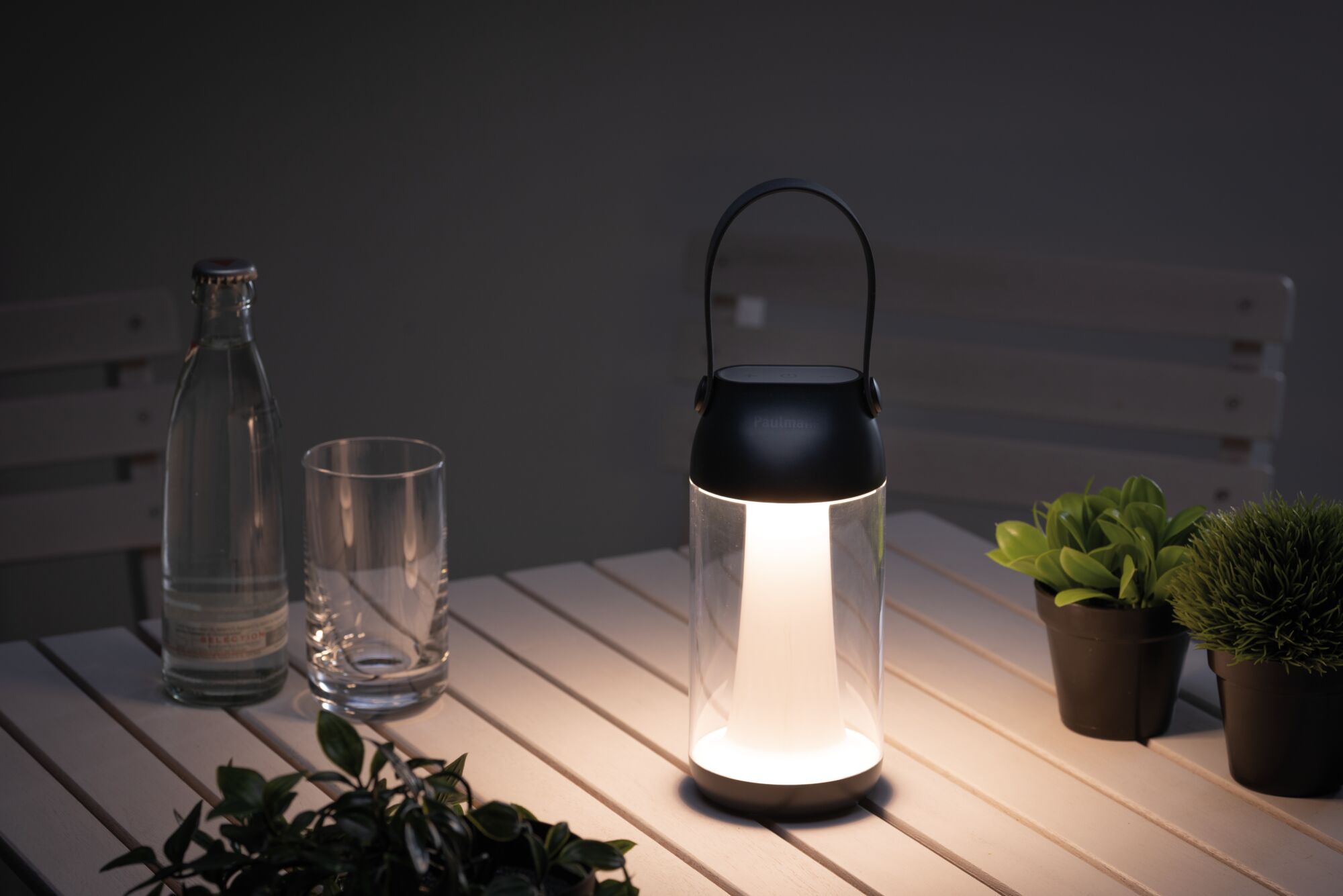 Moderne tragbare LED-Laterne in Schwarz mit warmweißem Licht auf weißem Gartentisch mit Pflanzen.