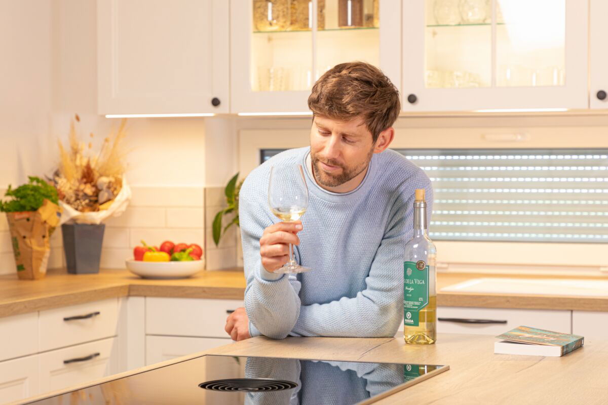 Man in blauwe trui proeft witte wijn in moderne keuken met houten aanrecht en fruitschaal