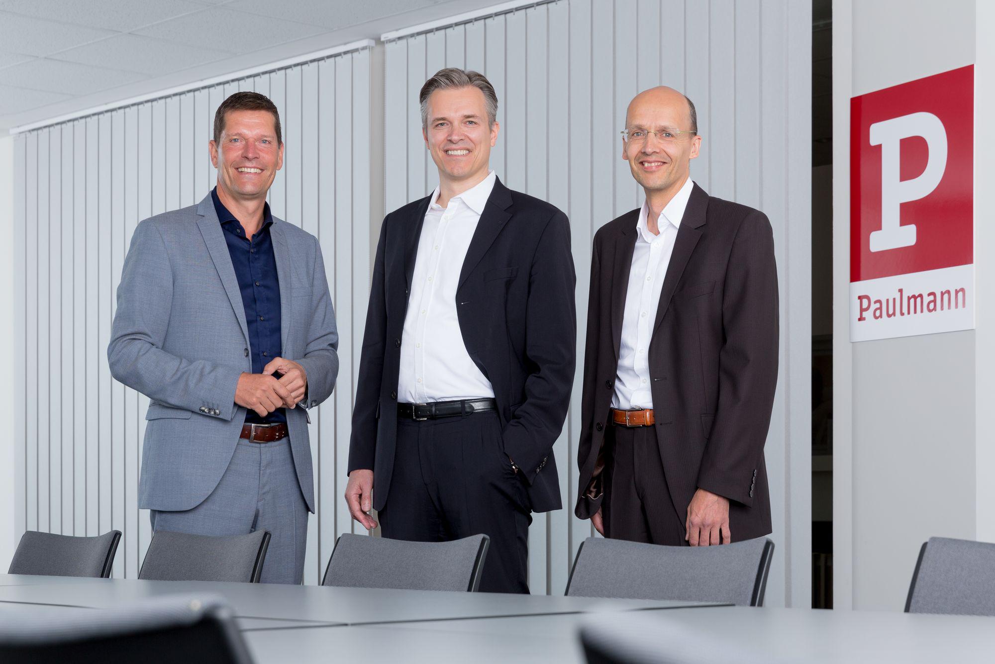 Three men in suits standing by Paulmann logo in modern office with conference table and chairs.
