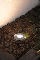 Round stainless steel recessed light with warm white LED in garden soil near green leaves.