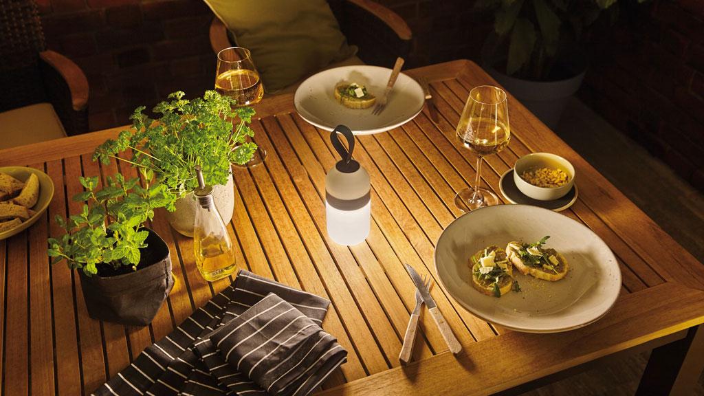 Gemütlicher Holztisch mit LED-Tischleuchte, Kräutertöpfen, Wein und kleinen Snacks für stimmungsvolle Beleuchtung.