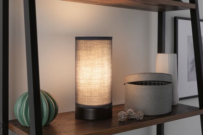 Cylindrical table lamp with light fabric shade and black base on wooden shelf with decor items.
