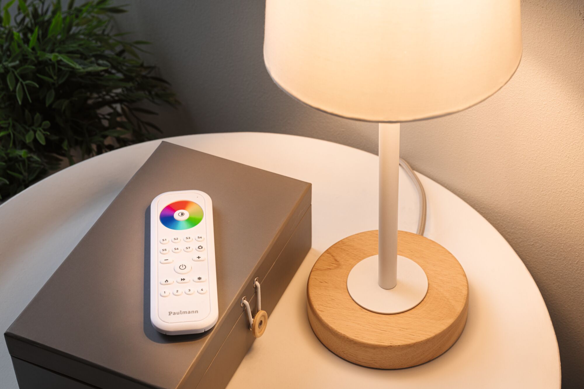 White remote with color wheel on gray box beside table lamp with wooden base on white table