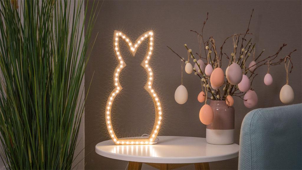 Lampe décorative LED en forme de lapin avec lumière blanche chaude sur table blanche près de branches de Pâques pastel.