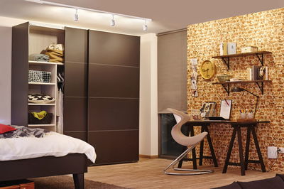 Modernes Schlafzimmer mit dunklem Holzkleiderschrank, Schreibtisch und Stuhl vor Ziegelwandbeleuchtung