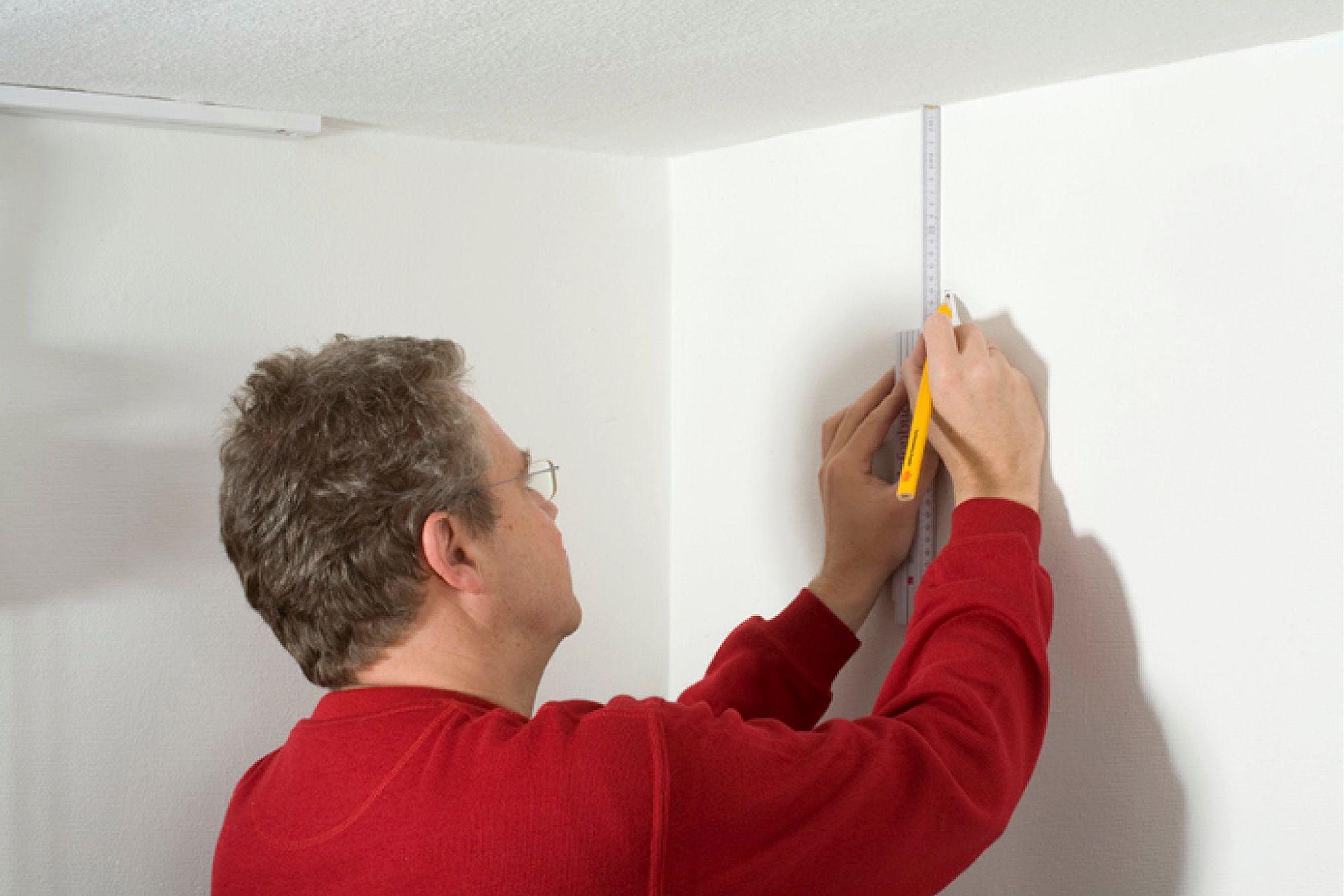 Mand i rød trøje måler hvid væg med lineal og blyant til belysningsinstallation.